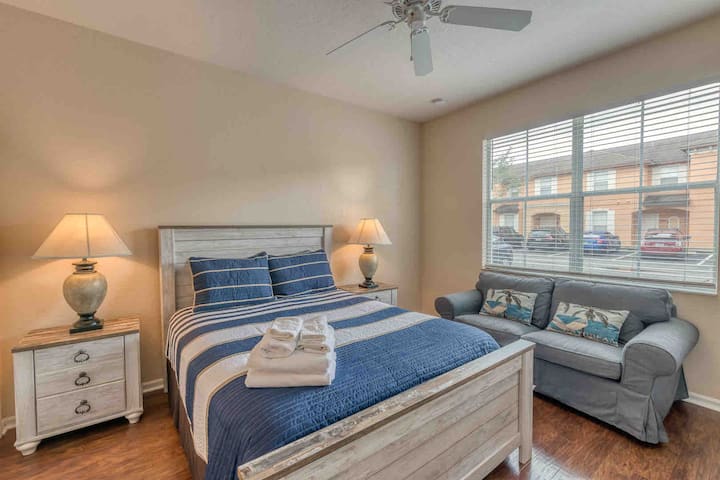 Chambre principale au rez-de-chaussée avec lit Queen Size, siège d'amour, télévision à écran plat, ventilateur au plafond et salle de bain attenante.