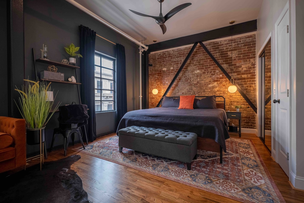 The bedroom features a spacious bed with a dark quilt and decorative pillows. A plush bench sits at the foot of the bed. The room is complemented by exposed brick walls, stylish shelving, and warm lighting from two pendant lamps.