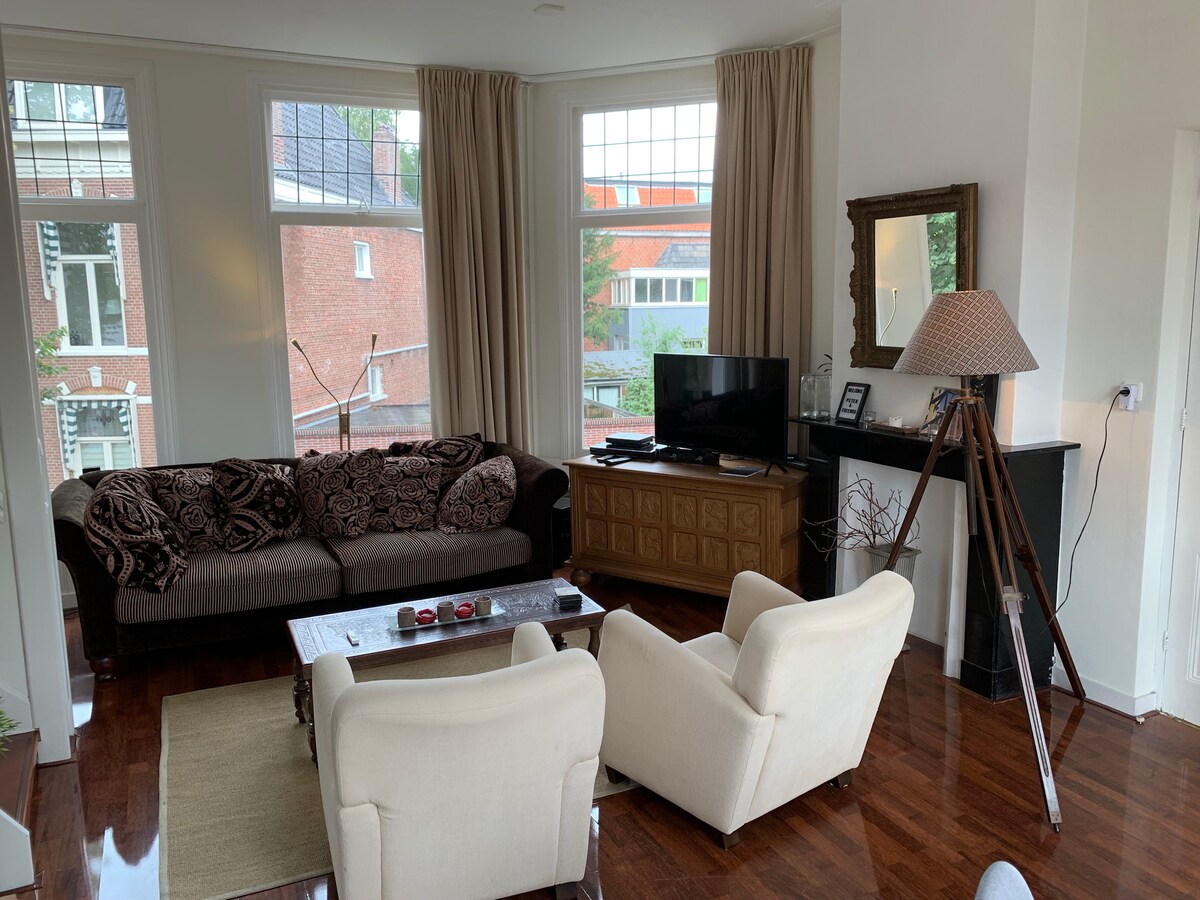 A spacious living room features a large couch adorned with patterned cushions and two comfortable armchairs. A wooden coffee table is centrally placed, and a television is positioned on a cabinet against the wall. Large windows allow natural light to brighten the space.