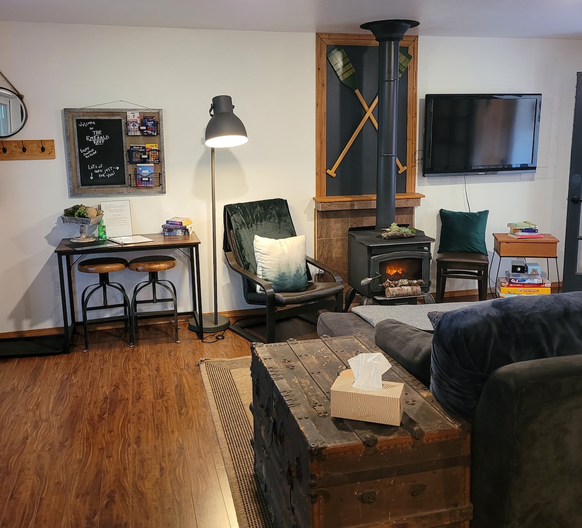A cozy living area is furnished with a green armchair and a black lamp beside a wooden table with bar stools. A simple entertainment unit displays a television above a modern gas stove, with rustic decor elements visible on the walls.
