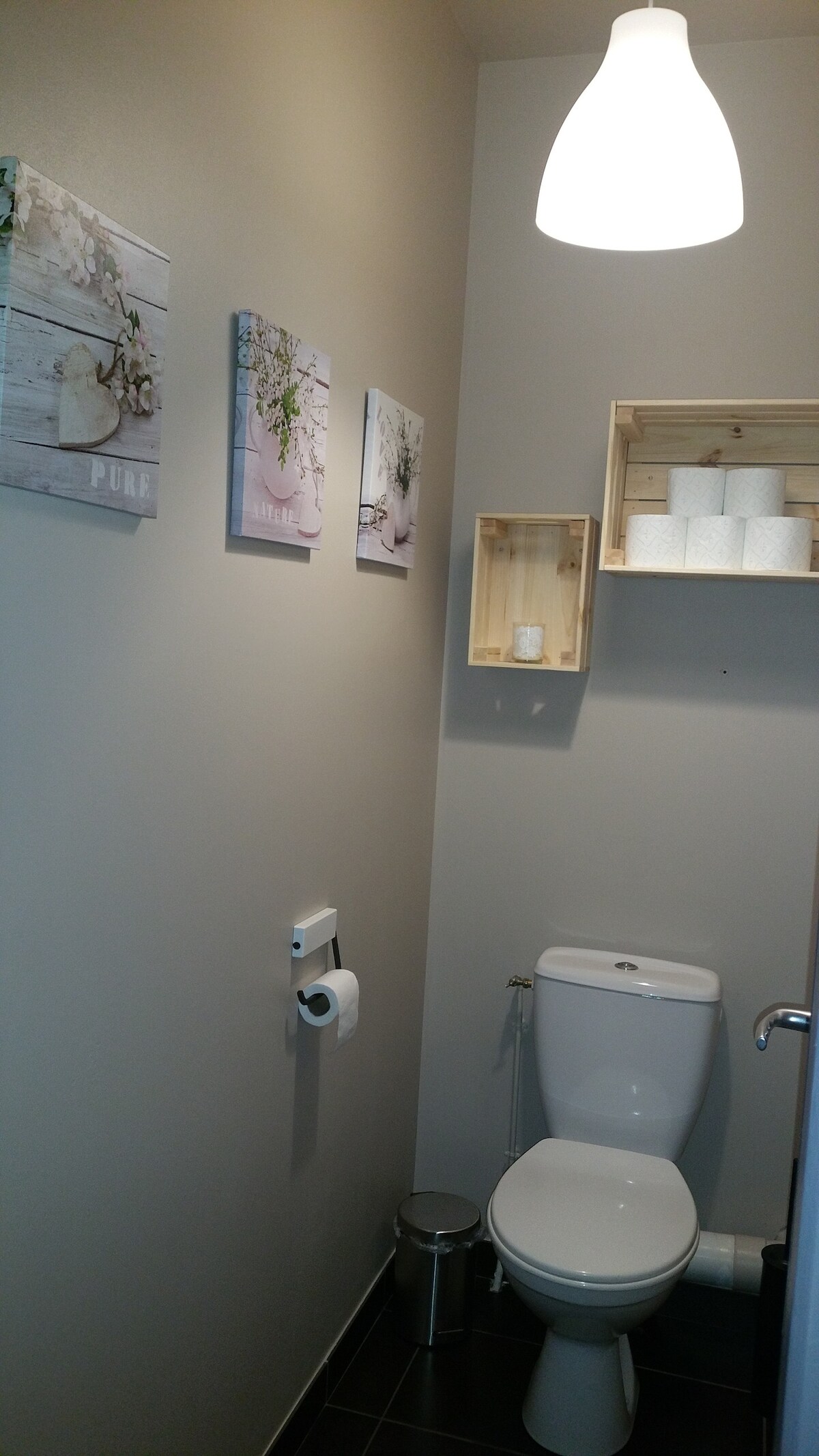 A clean and minimalistic bathroom is shown, featuring a white toilet and a small waste bin. Light wood shelving holds neatly stacked toilet paper rolls. Framed artwork adorned with floral themes enhances the neutral-colored walls. Soft lighting is provided by a pendant fixture above.