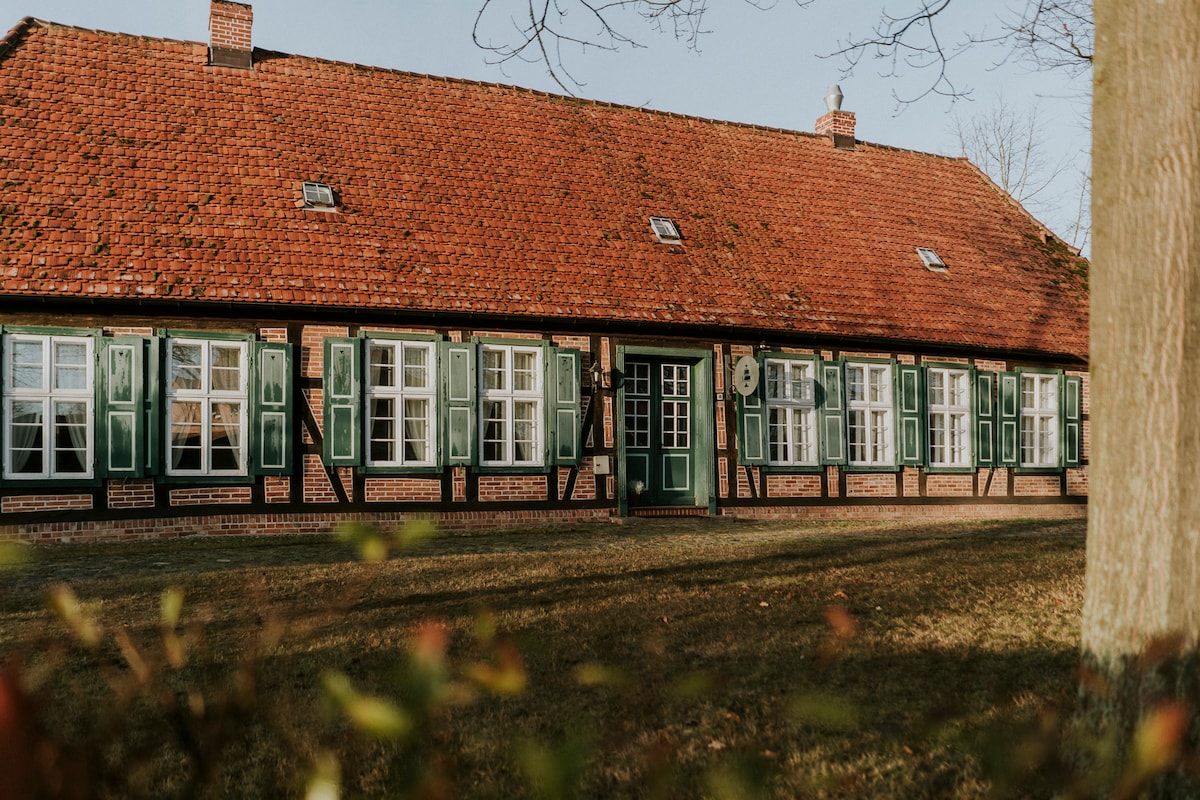 A traditional two-story building with a distinctive red-tiled roof and green shutters is featured. Large windows allow natural light to fill the interior. The structure is surrounded by a grassy area, creating a serene outdoor environment.