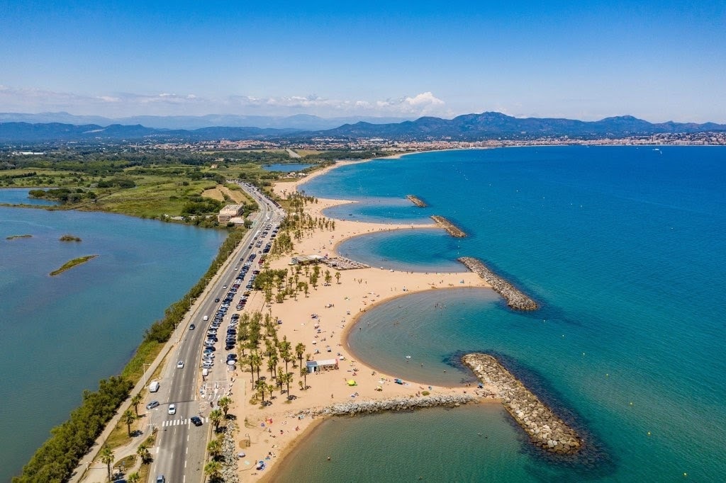 An aerial view captures a scenic beach coastline with gentle curves, bordered by a sandy shore and dotted with visitors. The sparkling blue water extends to distant hills under a clear sky, while a road runs parallel, lined with palm trees and vegetation.