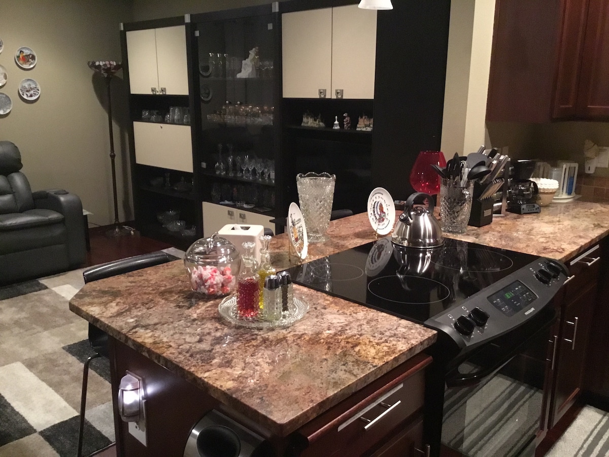 A modern kitchen space is presented, featuring a multi-toned granite countertop with a stove and various utensils. Glassware and decorative plates are displayed on a nearby cabinetry. A cozy seating area with a recliner chair is visible in the background.