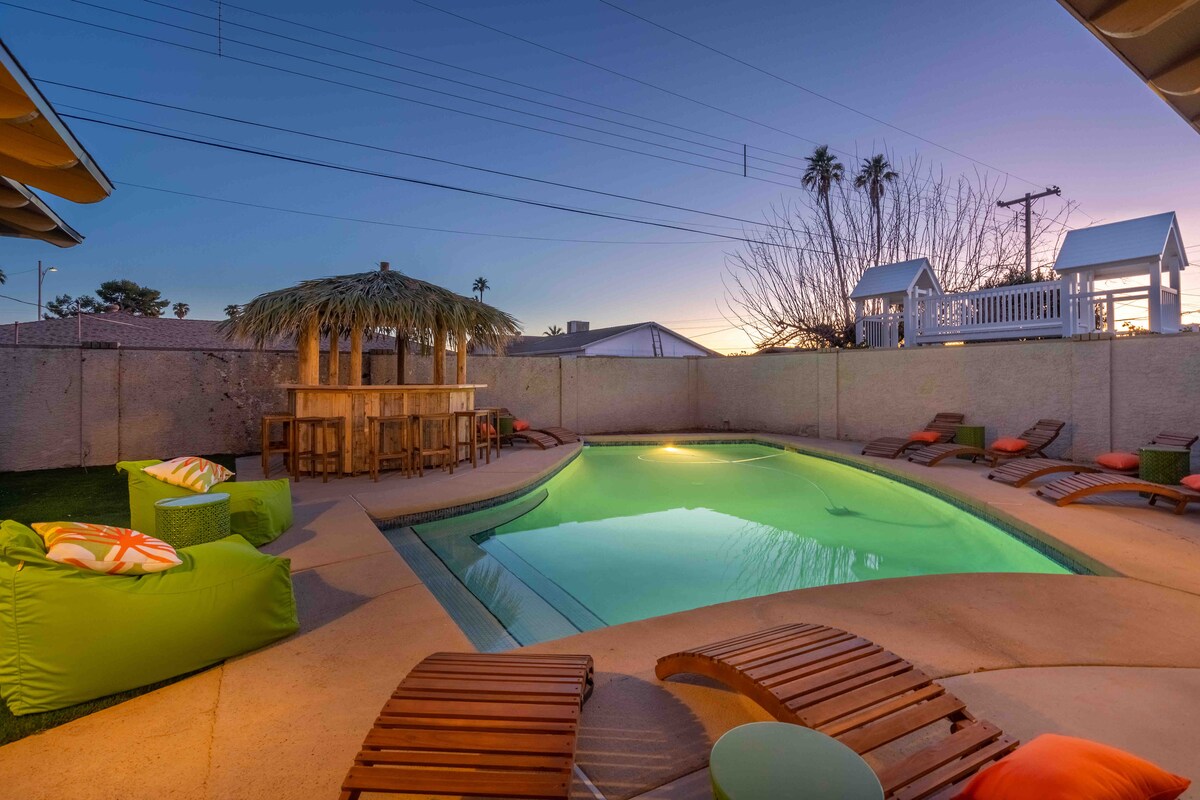 A heated pool is surrounded by a spacious patio, featuring vibrant lounge chairs and a tiki bar under a thatched roof. The serene water reflects the twilight sky, while palm trees and a play structure are visible in the background.