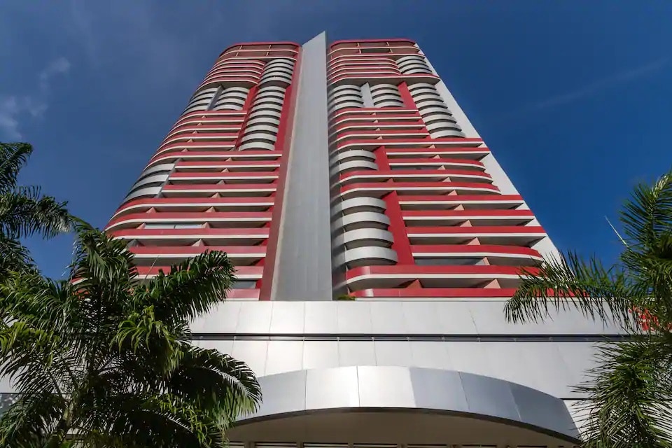 The modern high-rise building features a striking red and white facade, characterized by patterned balconies and expansive windows. Lush palm trees frame the entrance, enhancing the vibrant architectural design against a clear blue sky.