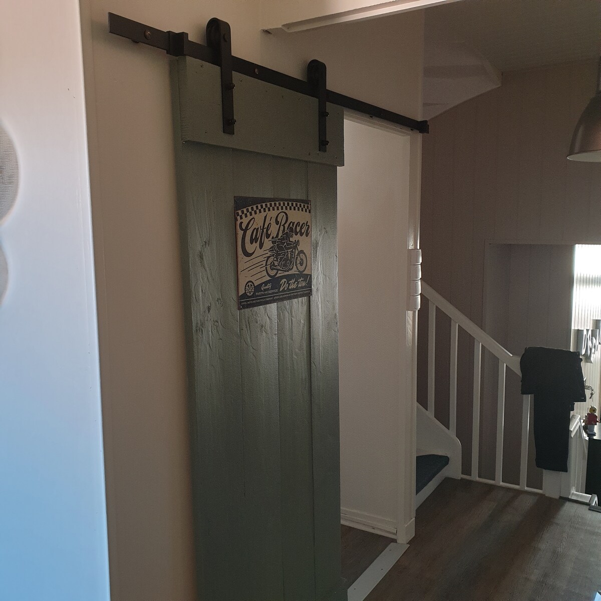 A sliding barn door, painted in green, features a vintage-style sign that reads 'Café Racer.' The door is framed by light-colored walls and adds a touch of character to the space. A staircase is visible in the background, leading to the upper level.