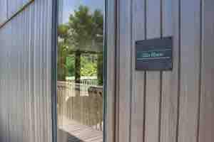 The entrance to Glas House is featured, showcasing a wooden exterior with a reflective glass door. A nameplate identifies the property, while green foliage is visible through the doorway, enhancing a connection to the surrounding nature.