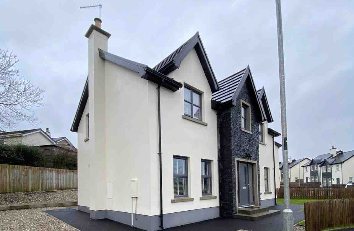 The modern two-story house features a mix of light and dark exterior walls, with a prominent peaked roof. Large windows are present on both levels, offering ample natural light. The well-maintained surrounding area includes a gravel driveway and wooden fencing, enhancing privacy.