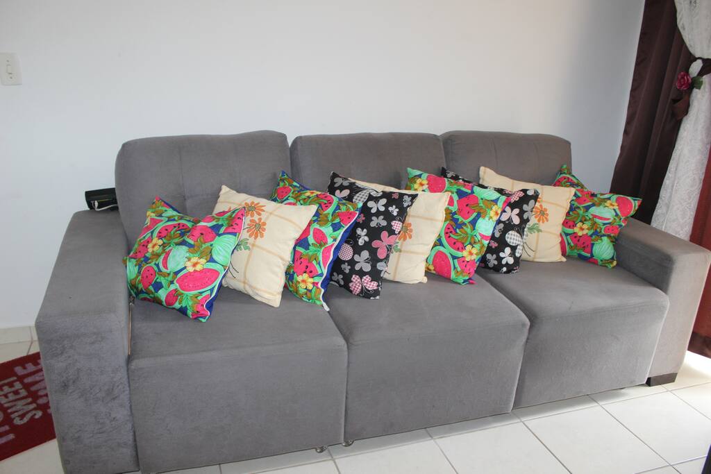 Sala de estar com televisão, sofá-cama retrátil de casal, cortinas, espaço para um colchão de solteiro e mesa de jantar.