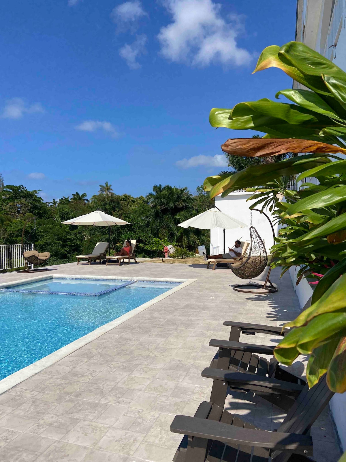 A clear blue pool invites relaxation, bordered by sun loungers and stylish umbrellas. Lush greenery provides a serene backdrop, while a nearby seating area features a hanging chair. The bright sky enhances the calm atmosphere of the outdoor space.