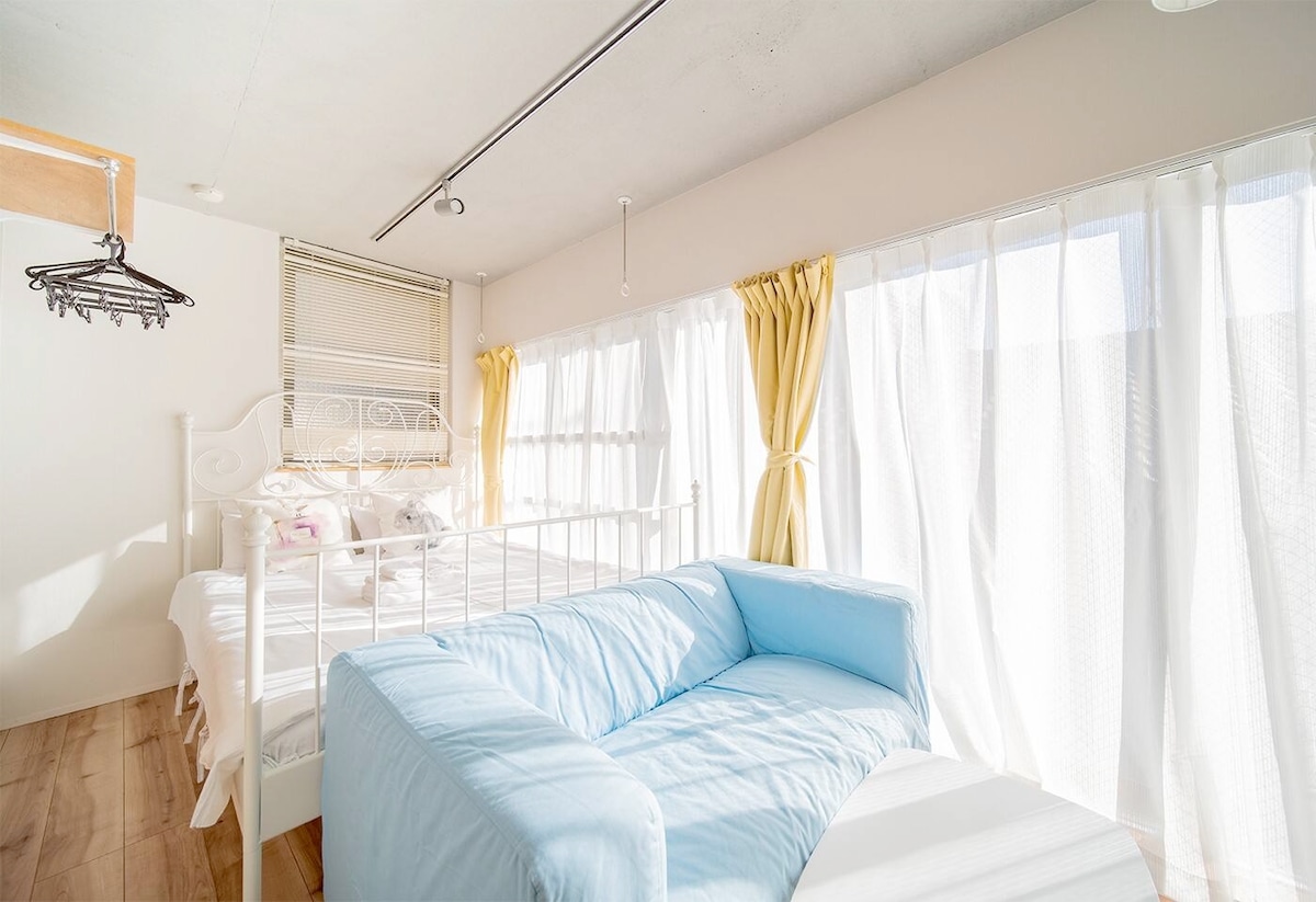 A sunlit room features large floor-to-ceiling windows with soft yellow curtains, providing ample natural light. A simple bed is positioned against one wall, accompanied by a light blue sofa in the seating area. The wooden floor adds warmth to the space.