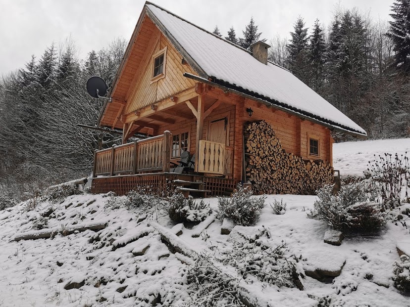 A wooden cabin is nestled in a snowy landscape, presenting a welcoming porch with wooden railings. Stacked firewood is arranged neatly beside the entrance, while snow-covered trees create a serene backdrop. A satellite dish is mounted on the roof, ensuring connectivity in a secluded setting.