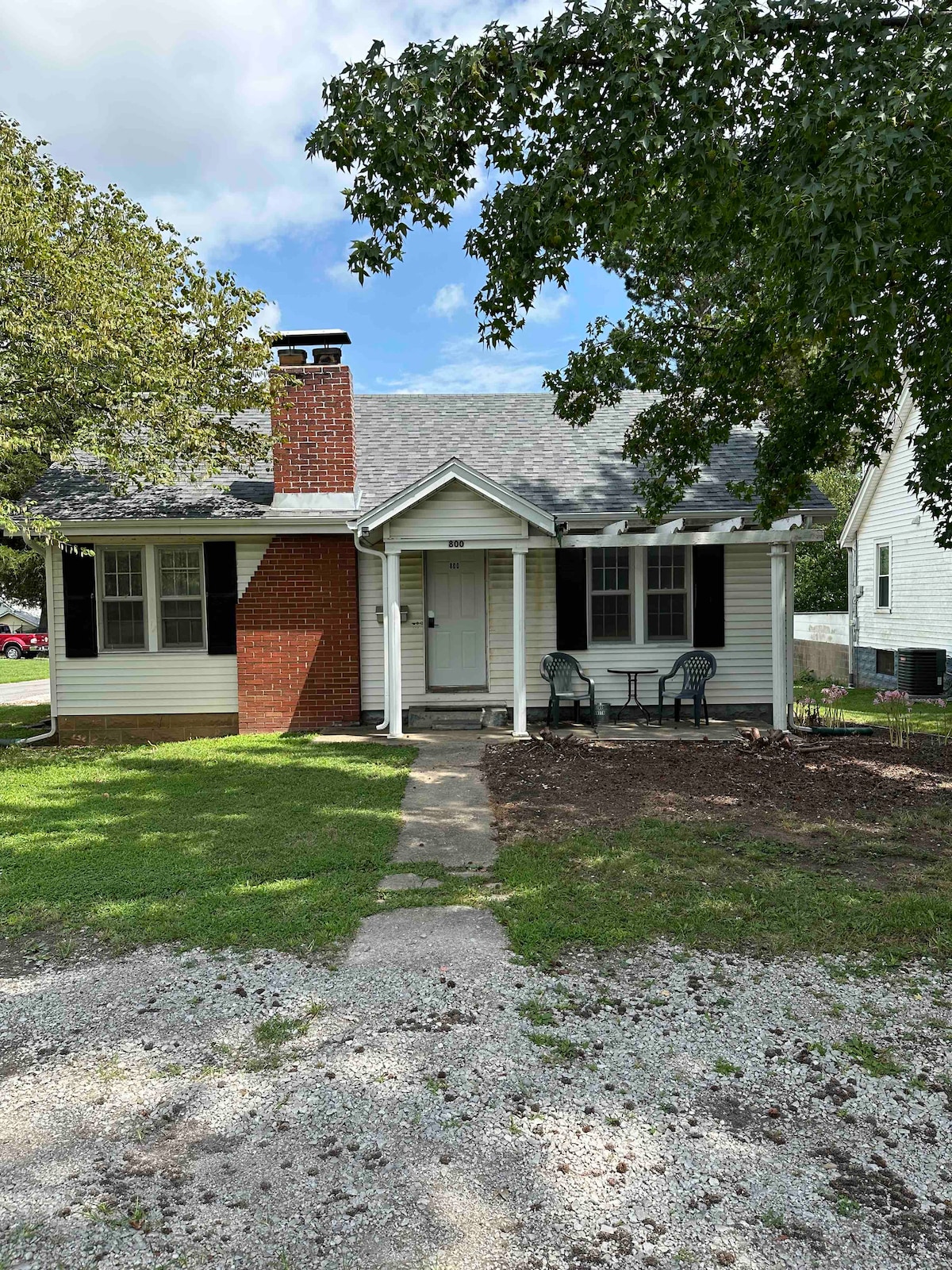 A vintage cottage is framed by lush greenery and a well-maintained lawn. A simple pathway leads to the front door, flanked by two chairs and a small table. The charming brick chimney adds character to the light-colored siding and dark shutters.
