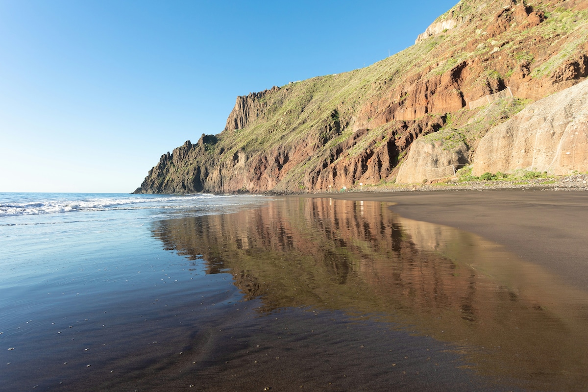 The image captures a serene beach scene with fine black sand reflecting the surrounding cliffs. Gentle waves lap against the shore, and the clear blue sky overhead enhances the tranquil atmosphere. The rugged cliffs are prominent in the background, showcasing earthy tones and vegetation.