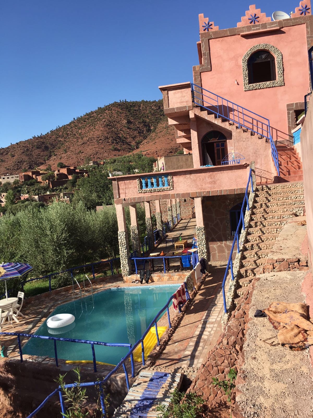 A traditional riad is displayed, set against a backdrop of rugged mountain terrain. A tranquil pool is situated beside the building, surrounded by olive trees and gardens. Steps lead up to the upper level, while colorful railings add character to the exterior.