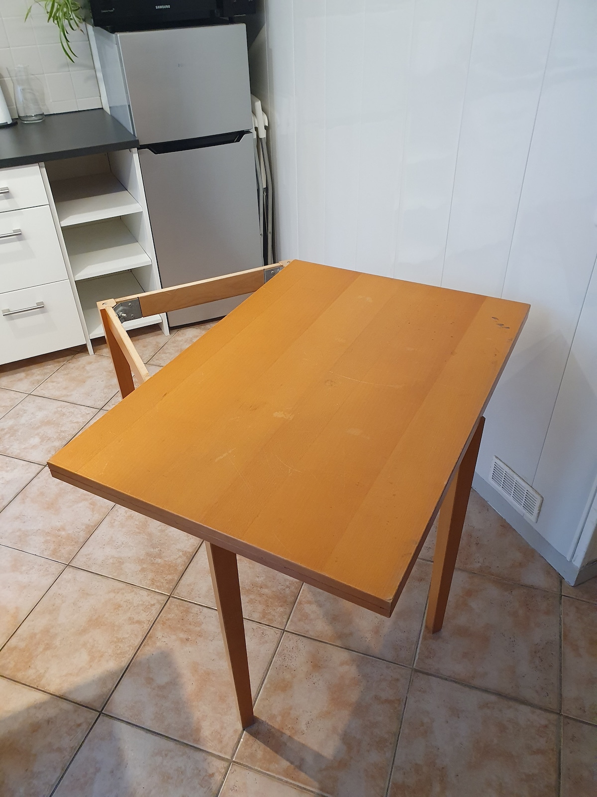 A wooden table with a rectangular top and two legs is positioned in a kitchen area. One side of the table is partially detached, indicating a potential foldable design. The tiled floor and cabinetry in the background are visible, contributing to the functional kitchen setting.