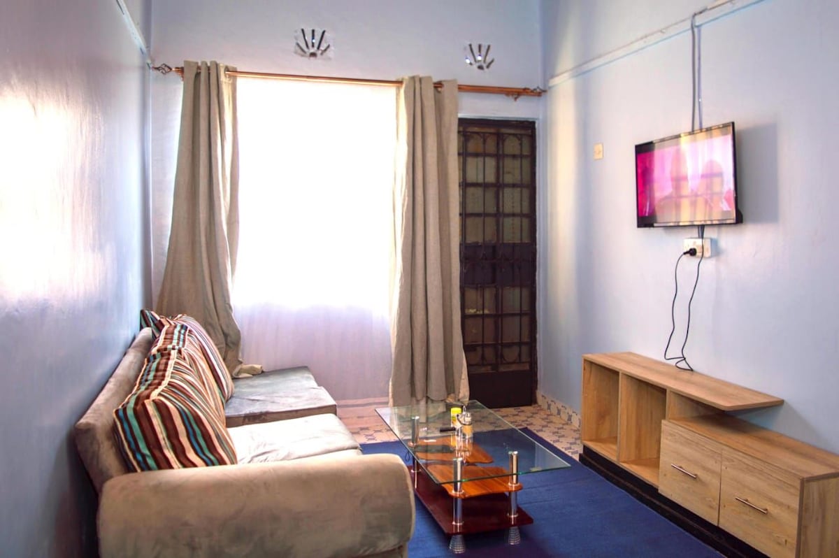 A light-filled living area features blue walls and a combination of a sofa and glass coffee table. A television is mounted on the wall opposite the seating area. Sheer curtains frame the window, while a wooden cabinet is positioned nearby.