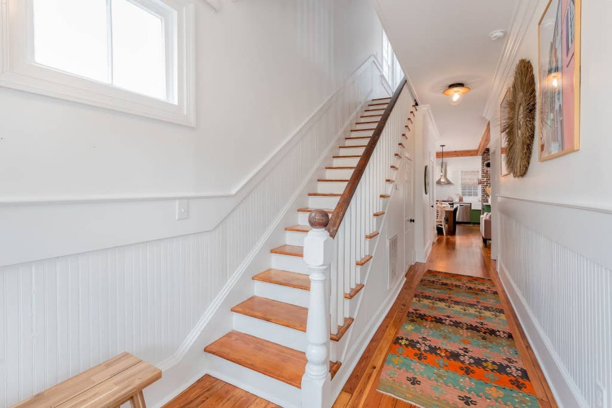 A light-filled hallway features a wooden staircase leading to the upper level. The walls are painted white, creating a fresh ambiance. A colorful patterned rug runs along the wooden floor, and a bench is positioned to the side, adding functionality to the space.