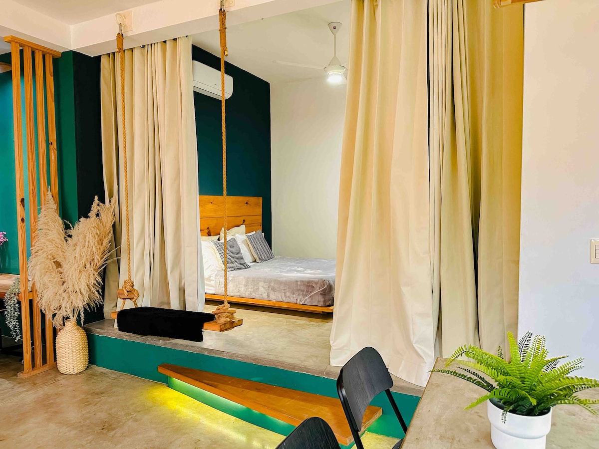 An open space features a division created by flowing curtains, leading to a cozy sleeping area with a wooden bed. Soft lighting highlights the green accents and the concrete floor. A swing adds a playful touch, while a potted plant enhances the natural feel.