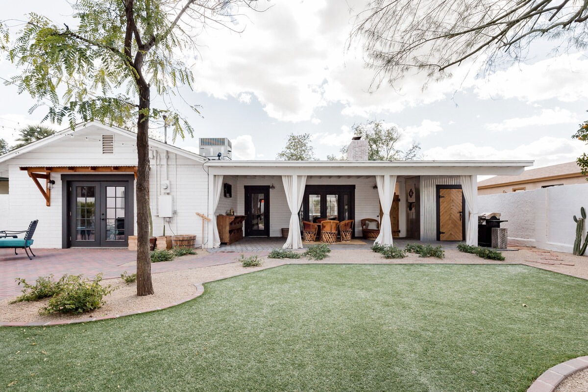 The exterior showcases a spacious backyard featuring a well-maintained lawn surrounded by desert landscaping. A covered patio area offers seating options and a dining space beneath fabric drapes, with a stone wall and greenery enhancing the ambiance.