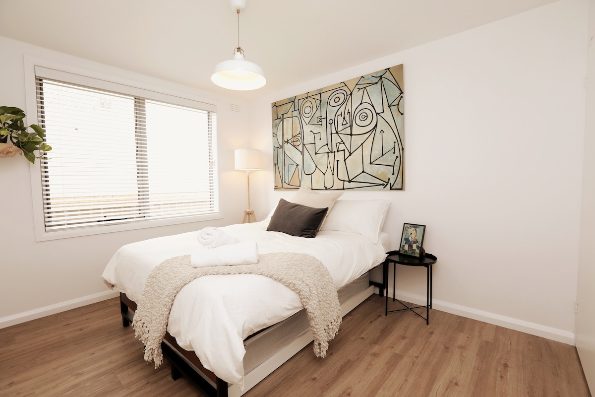 A well-lit bedroom features a spacious bed covered with quality linens and a cozy throw. A piece of abstract wall art hangs above the bed. Natural light enters through a window adorned with blinds, and a small bedside table holds a framed picture.