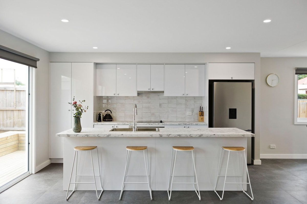 A modern kitchen showcases a marble-topped island bar with four stools. Sleek white cabinetry and a stylish backsplash complement the appliances, while natural light filters through the window. A vase with fresh flowers adds a touch of color to the neutral palette.