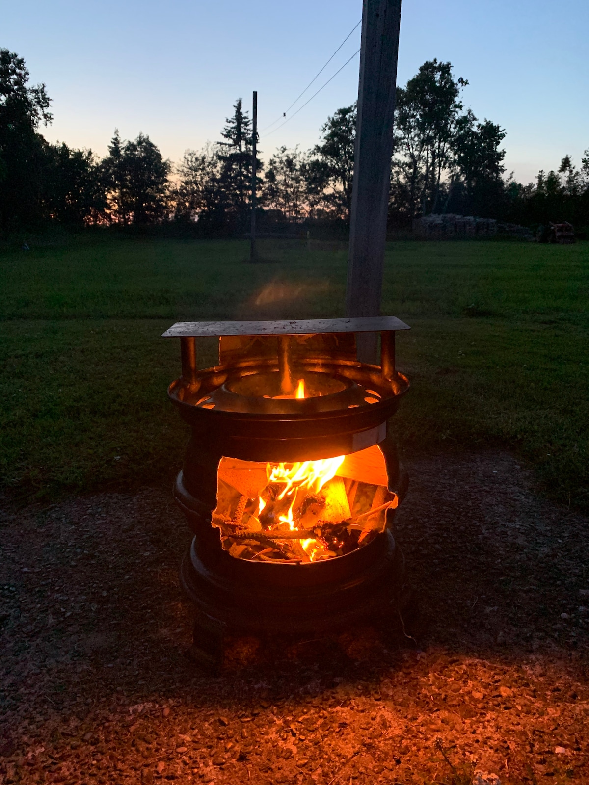 A fire pit constructed from a metal drum showcases a vibrant flame at dusk, with smoke gently rising into the evening air. The surrounding grassy area is illuminated by the warm glow of the fire, offering a cozy ambiance in the open countryside.