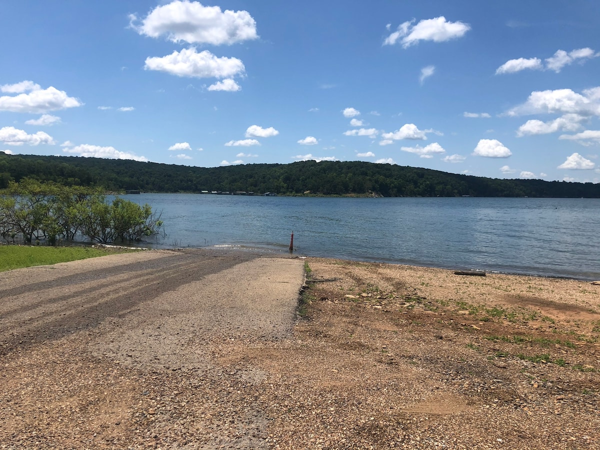 A serene view of Lake Tenkiller features a gentle shoreline with a gravel access point leading into the water. Lush green hills rise in the background under a bright blue sky dotted with fluffy white clouds.