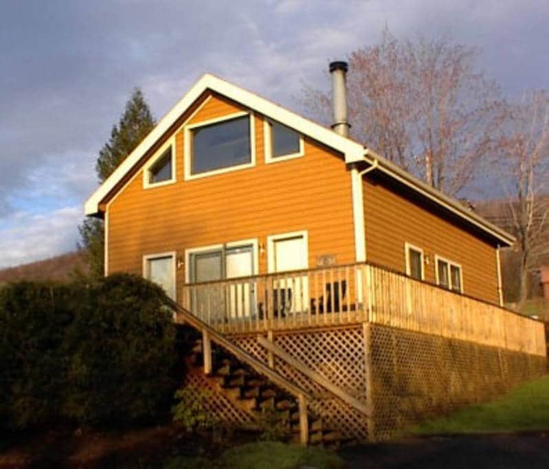 The chalet features a warm wood exterior with large windows, allowing natural light to fill the interior. A spacious front deck is accessible via stairs, offering a welcoming entrance and space for outdoor seating. Lush greenery surrounds the property.