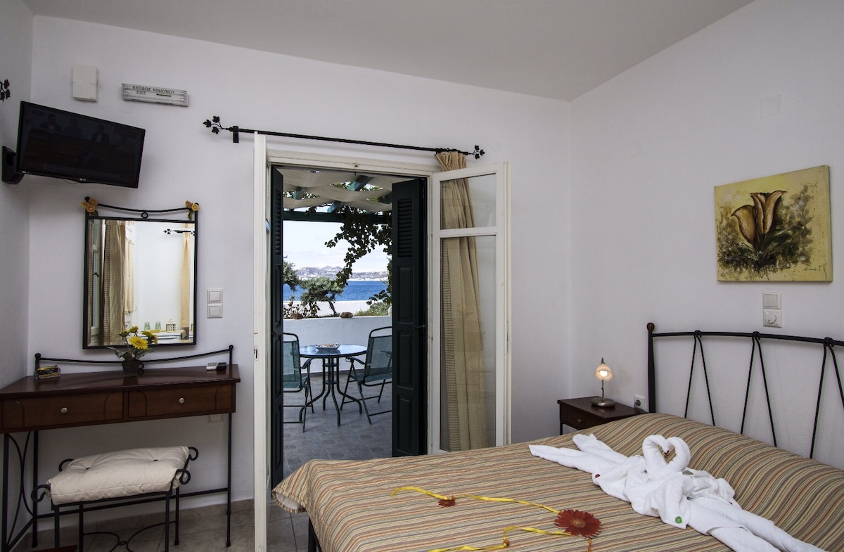 The room features a double bed with a striped blanket and decorative towels. A console table with a mirror is situated beside the bed, complemented by a small lamp. The balcony doors open to reveal outdoor seating with a sea view.