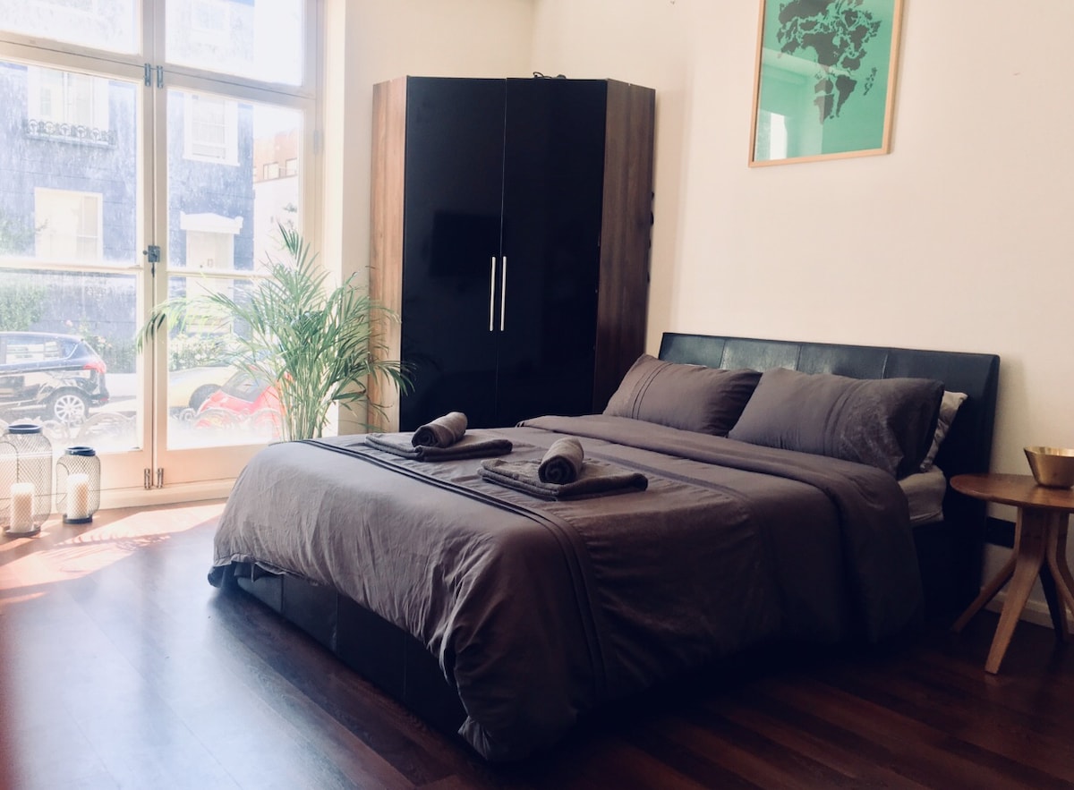 A spacious sleeping area is presented with a large bed dressed in neutral-toned bedding, complemented by two towels neatly arranged. A wardrobe in dark wood stands against the wall, while a plant adds a touch of greenery. Natural light floods in from the large windows.