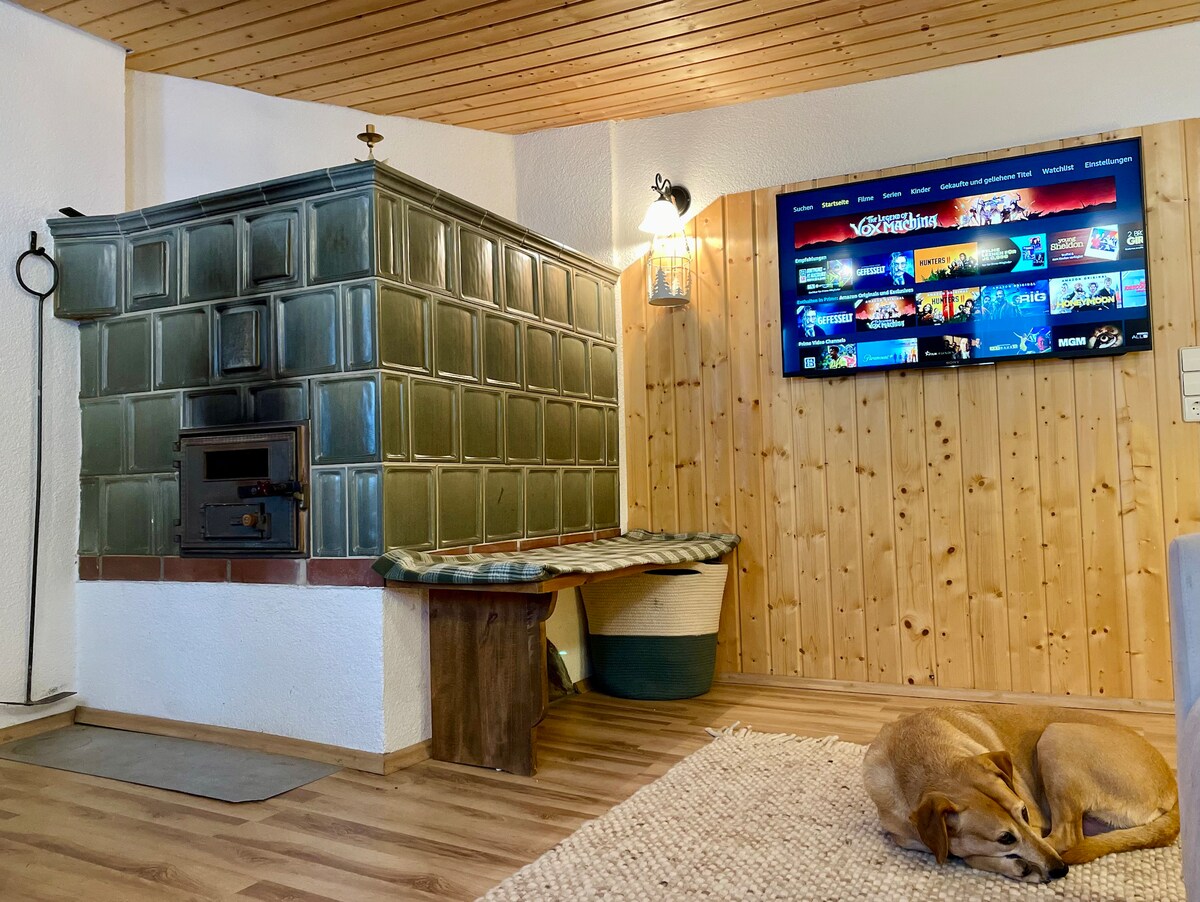 A tiled stove is featured in the living area, providing a rustic focal point. A cozy seating nook is positioned beside it, and a modern television mounted on the wall displays various media options. A dog rests comfortably on the floor, adding a homey touch.
