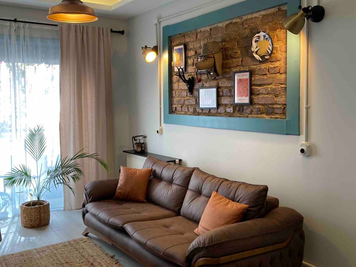 A cozy living space features a leather sofa adorned with cushions in warm tones. A decorative wall with exposed brick showcases framed art pieces. Soft light is provided by industrial-style lamps, while a floor plant adds a touch of greenery to the welcoming atmosphere.