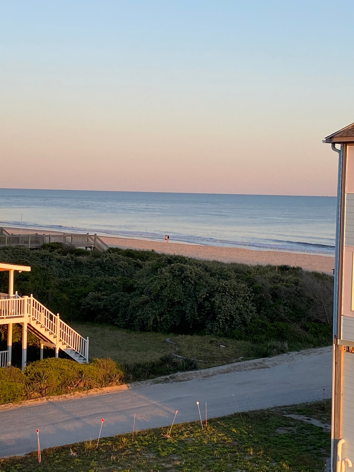 The image captures a serene beach scene during sunset, with gentle waves lapping against the shoreline. A lone figure can be seen walking along the water's edge, while lush greenery and sand dunes frame the beach. The sky displays soft pastel hues, enhancing the tranquil atmosphere.