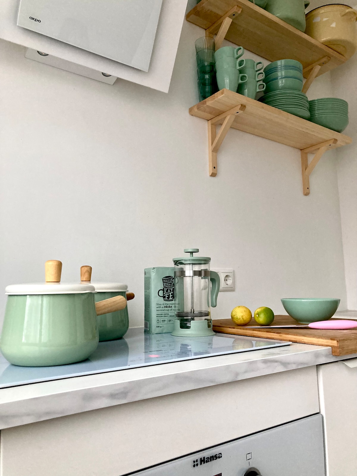 A modern kitchen area features light green cookware and a French press on a marble countertop. Above, open shelves display a collection of stacked plates and mugs. A wooden cutting board and a bowl are positioned nearby, accompanied by fresh limes, enhancing the inviting setup.