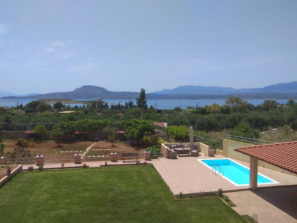 The view features a serene landscape with a clear blue pool adjacent to a well-maintained lawn. In the background, mountains and trees frame the scene, while the coastline outlines the horizon. Sunlight illuminates the area, enhancing the tranquil outdoor setting.