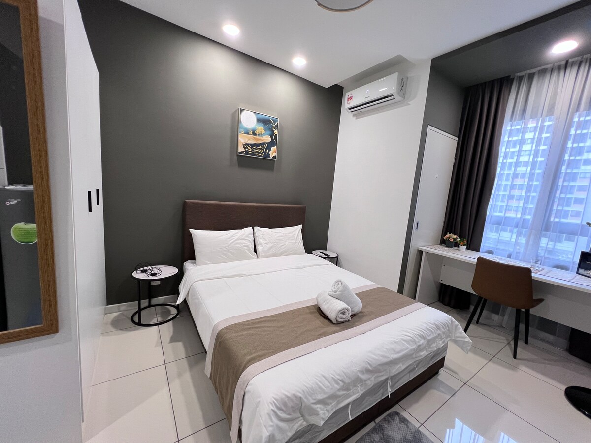 The room features a queen-sized bed with a neutral-colored blanket and two pillows. A workspace with a chair is present beside the bed, with a small table on either side. Large windows provide natural light, while air conditioning maintains a comfortable environment.
