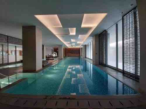 An indoor swimming pool features a clear blue water surface, illuminated by soft overhead lighting. Floor-to-ceiling windows on one side allow natural light to fill the space, while sleek tiled walls provide a modern aesthetic. Lounge chairs are arranged beside the pool.