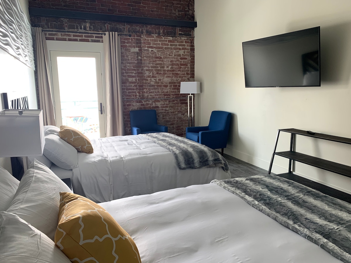 A well-lit guest room features two spacious beds dressed in white linens, accented by a decorative yellow pillow. A flat-panel television is mounted on the wall, while a seating area with two blue chairs is positioned near a glass door leading to a balcony.