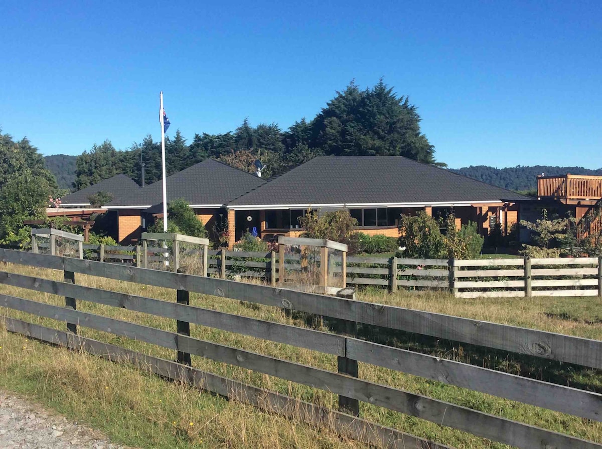 The exterior of a spacious country home is viewed from a distance, surrounded by lush greenery and rolling hills. A wooden fence lines the front, complementing the neatly maintained garden. A flagpole is positioned prominently, with the house featuring a dark roof and large windows.