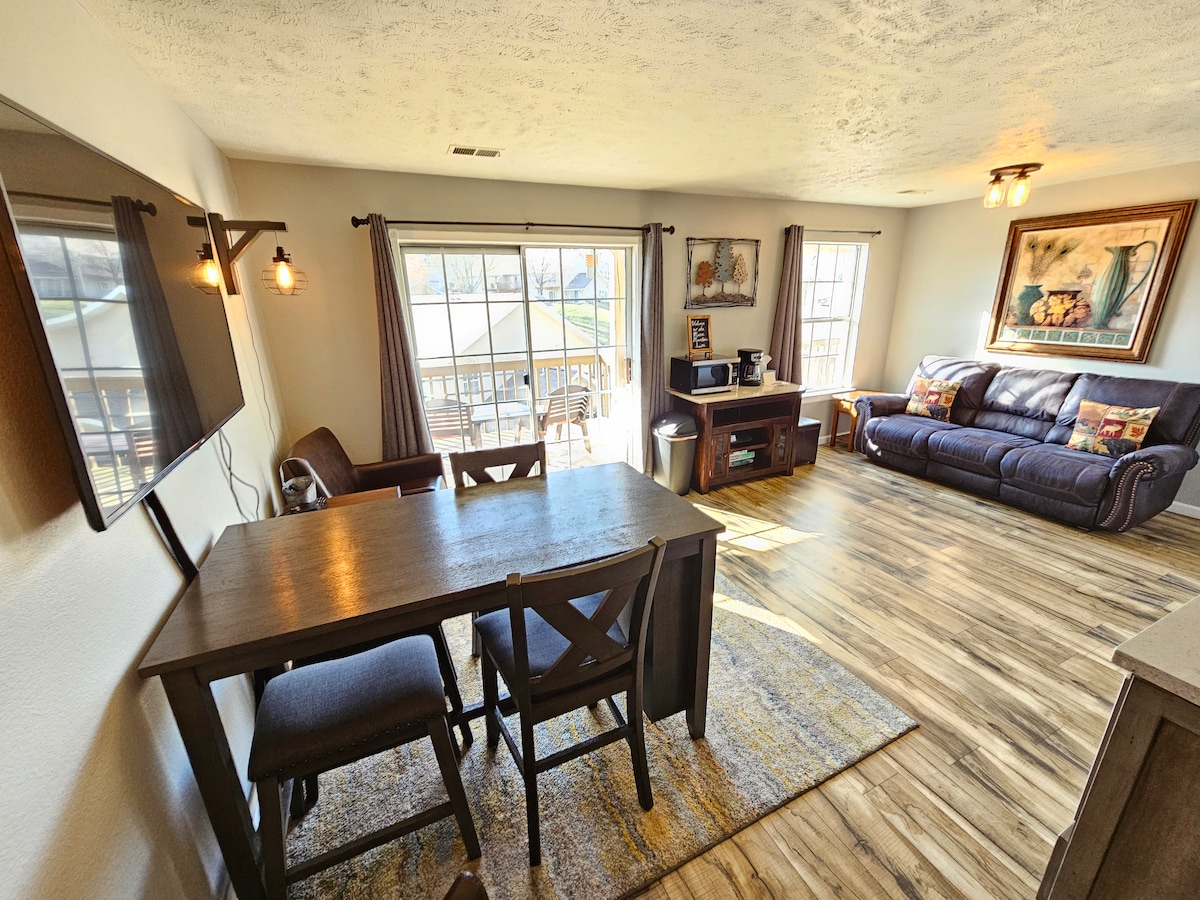 A spacious living area features large windows that allow for natural light. A comfortable couch is positioned beside a wooden entertainment unit and a smart TV. A dining table with chairs stands nearby, complemented by a neutral rug that adds warmth to the space.