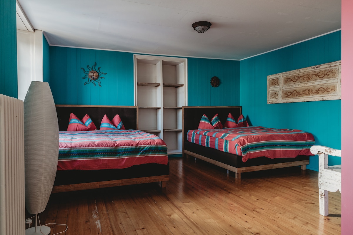 A brightly colored room features two spacious beds with striped bedding, complemented by decorative pillows. The walls are painted in a vibrant blue, and a decorative sun motif adds a playful touch. Shelving units are visible, providing additional storage options.