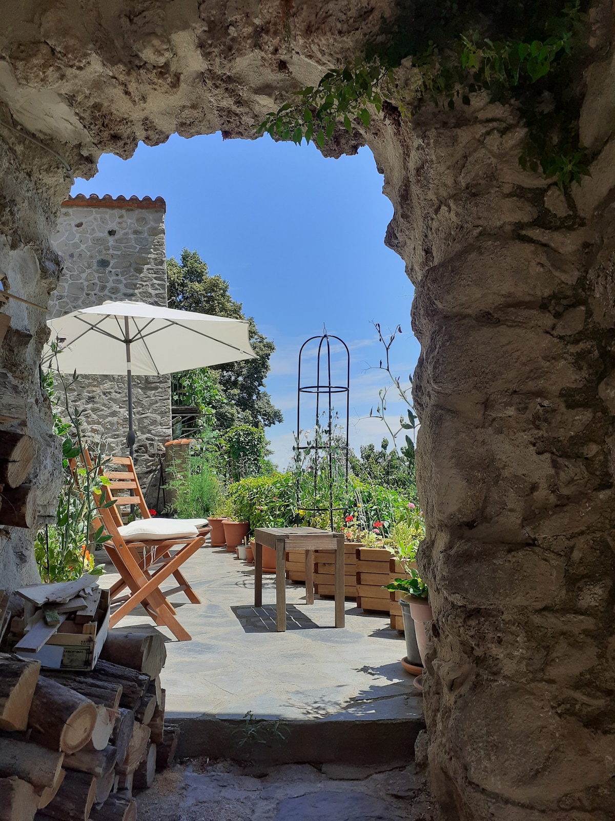 An arched stone doorway frames a calm outdoor space. Two wooden chairs are positioned next to a small table, with an umbrella providing shade. Lush greenery and potted plants are visible in the background, enhancing the serene atmosphere of the private terrace.