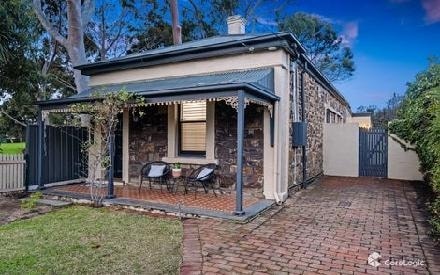 The exterior features a charming stone facade and a covered porch with two chairs. A paved path leads to a private gated entryway. Lush greenery surrounds the area, enhancing the serene setting near a park.