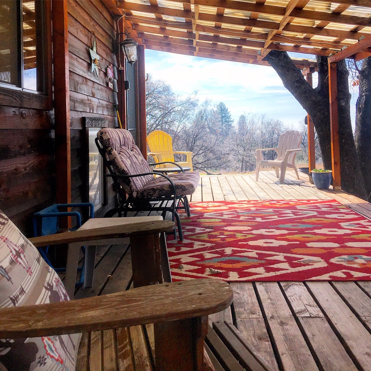 A wooden deck featuring a patterned rug and a mix of seated areas, including a rocking chair and colorful outdoor chairs. Sunlight filters through a canopy, illuminating the surrounding trees and creating a serene outdoor space. Nature is visible in the background, enhancing the setting.