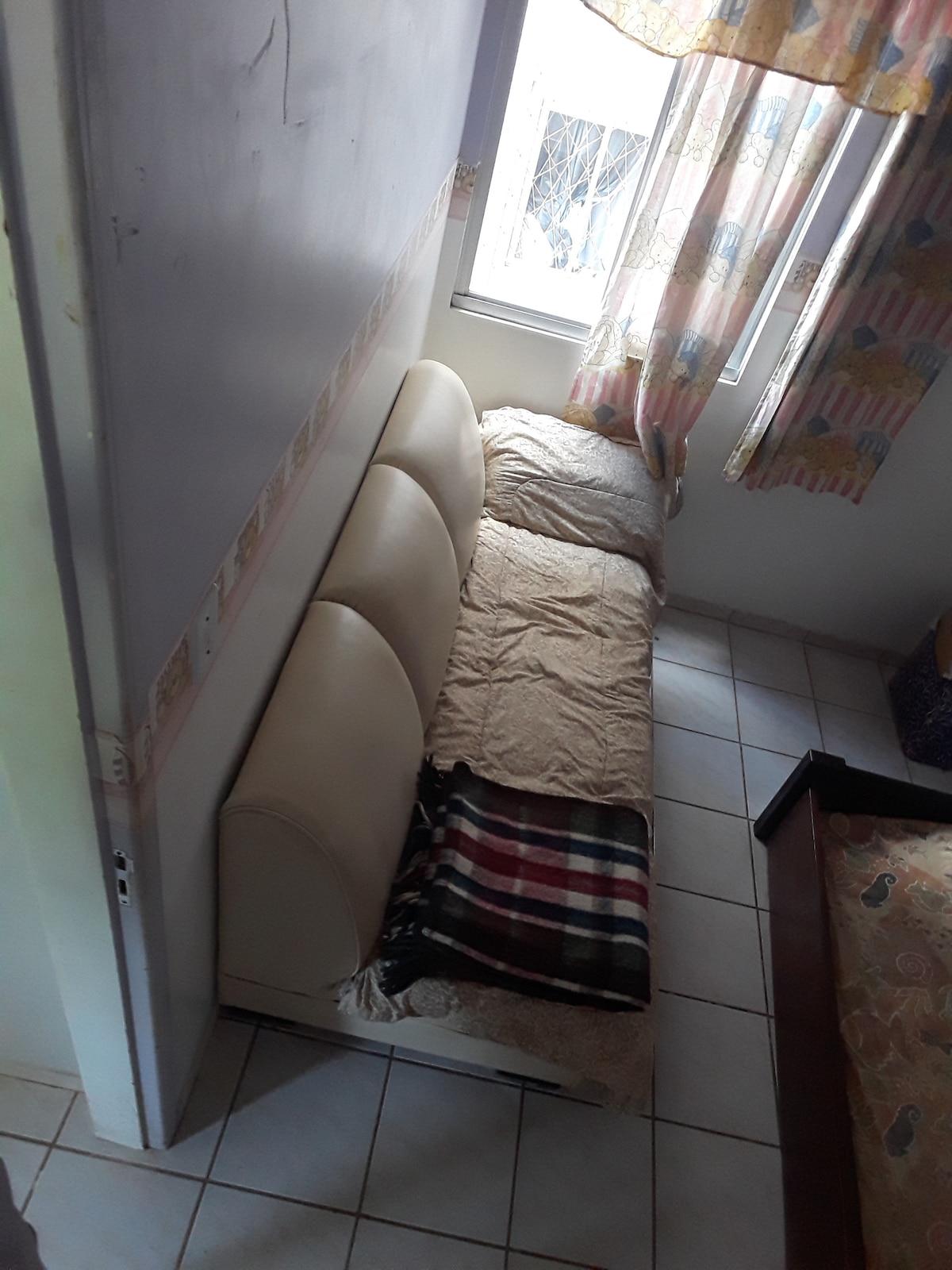 A compact seating area features a light-colored, three-cushion sofa covered with a beige throw. A plaid blanket is casually draped over the sofa. Natural light enters through a nearby window adorned with patterned curtains.