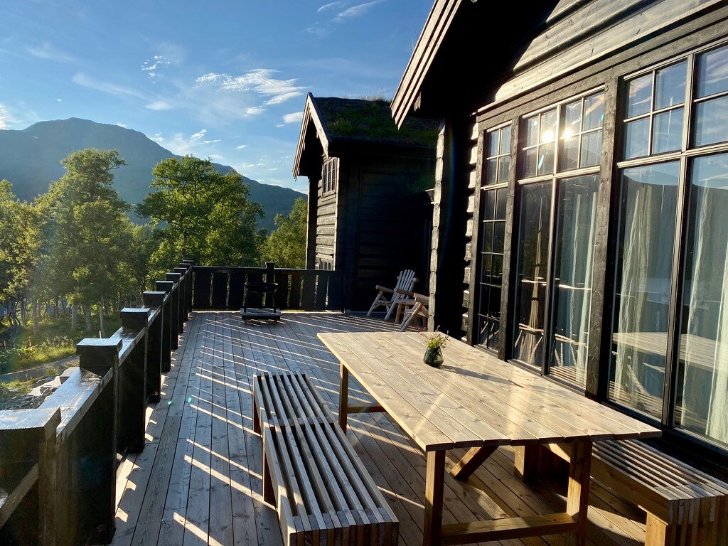 A spacious deck is featured, surrounded by trees and with mountain views in the background. A large wooden table is positioned at the center, accompanied by several benches. Large windows reflect natural light, enhancing the connection between indoor and outdoor spaces.