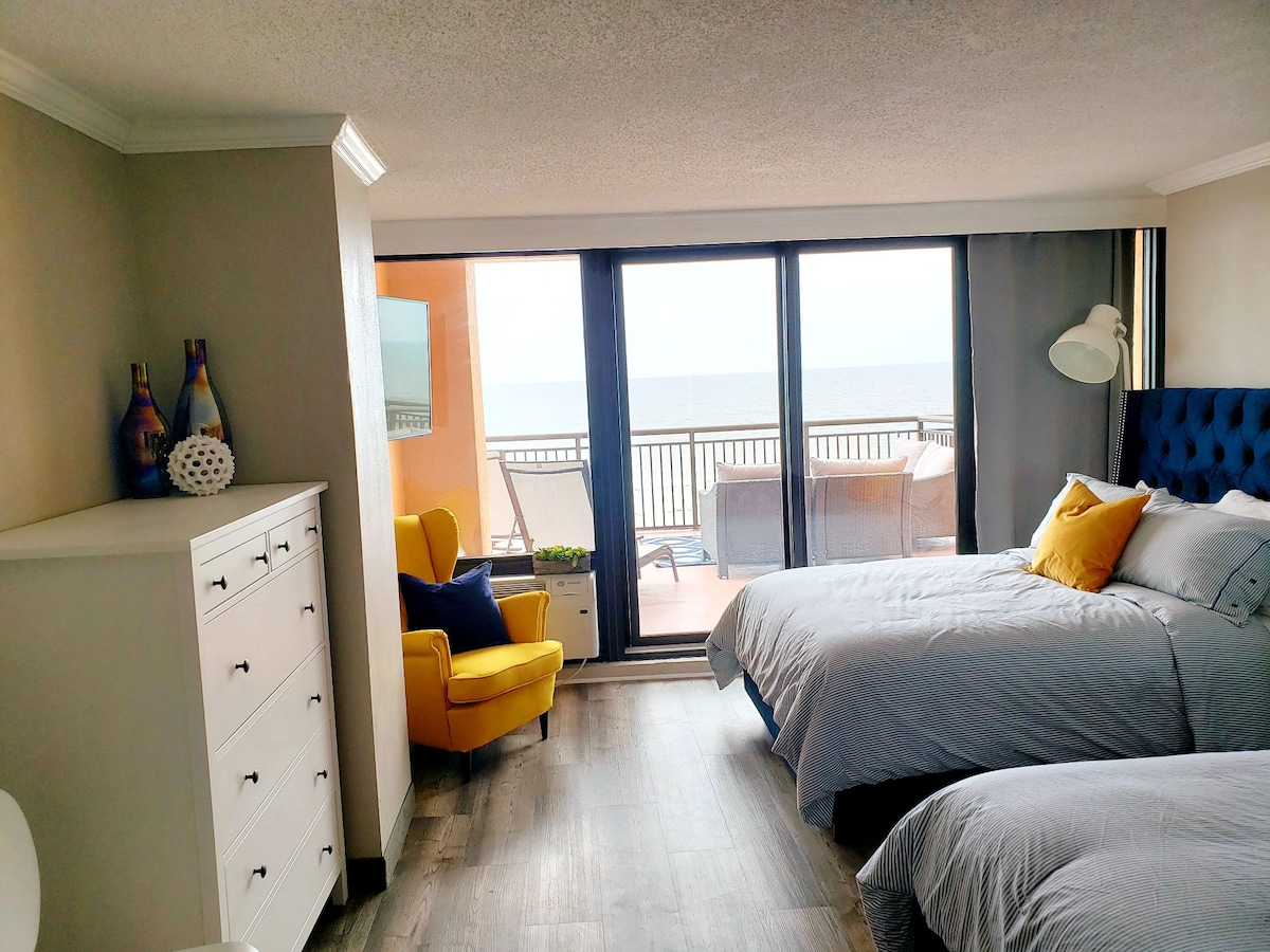 The bedroom contains two full size beds with soft linens, a bright yellow chair, and a dresser. Large glass doors lead to a private terrace with ocean views, allowing natural light to illuminate the space.