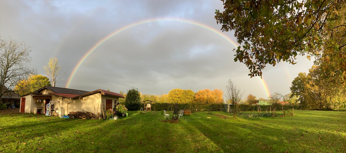 A wide grassy area is framed by trees, with a rainbow arcing across a cloudy sky. A single-story building with a rustic exterior is visible, alongside garden features such as chairs. Colorful autumn foliage can be seen in the background.